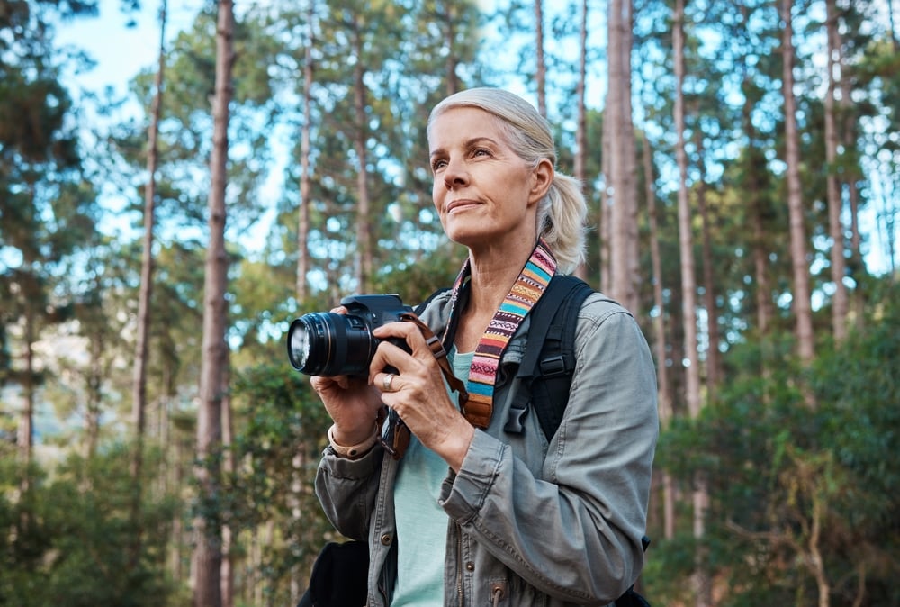 Woman with camera stands in forest, wearing a backpack and colorful strap.