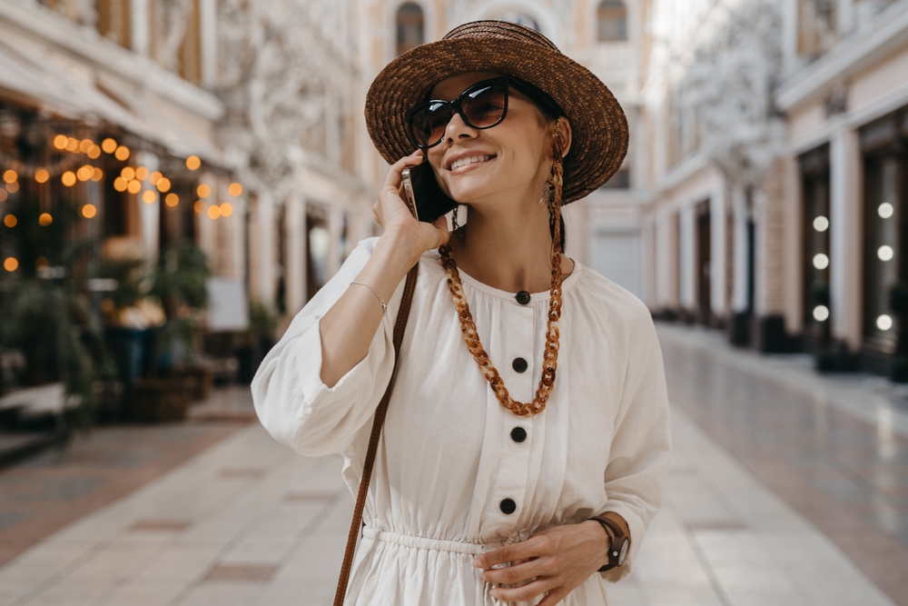 A woman in a white dress and hat, talking on a phone, standing in an outdoor plaza.