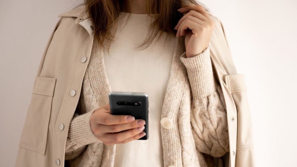 A person in a beige coat and sweater holds a smartphone, with only torso and hand visible.