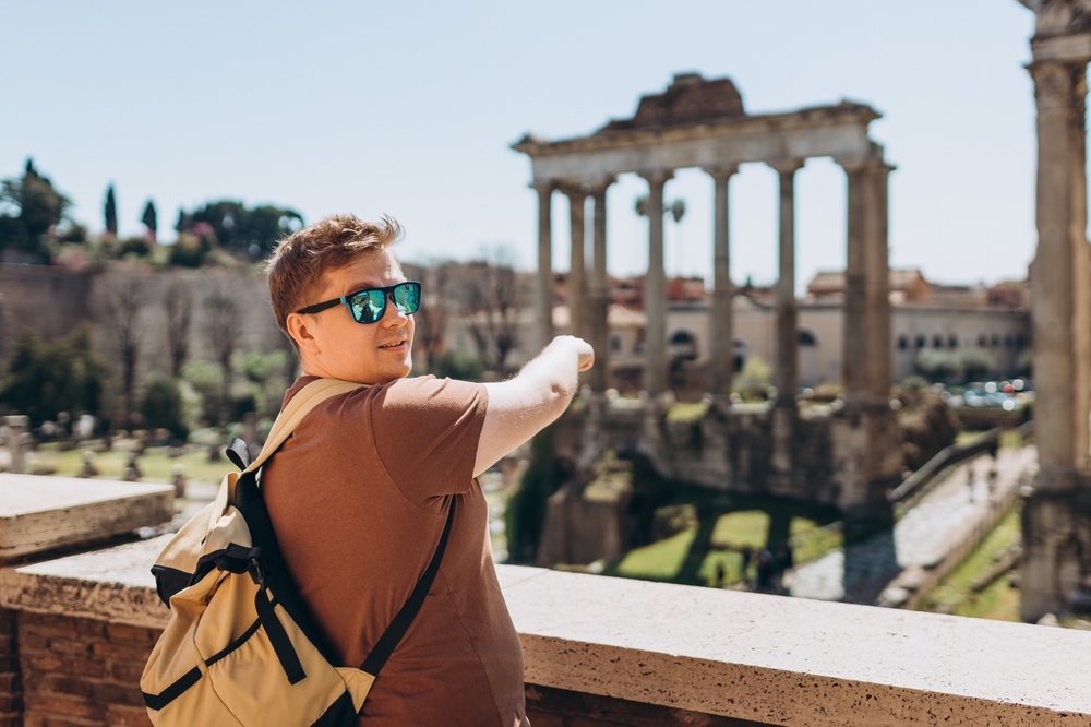 A person with sunglasses and backpack pointing to ancient ruins with columns in the background.