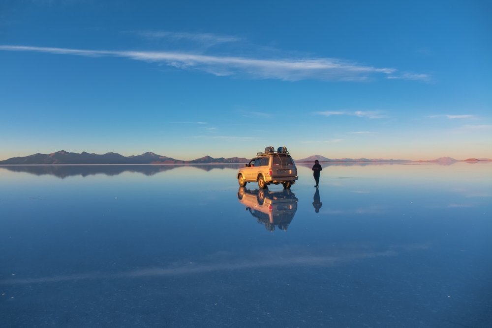 SUV and person on reflective salt flat under clear blue sky at sunset.