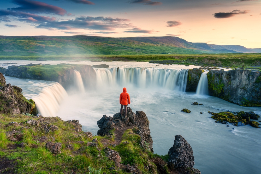 Person in orange jacket stands on rocks, overlooking a wide waterfall and lush landscape at sunset.