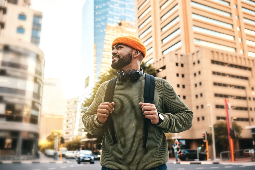 Man in green sweater, orange beanie, with headphones and backpack, standing on city street with tall buildings.