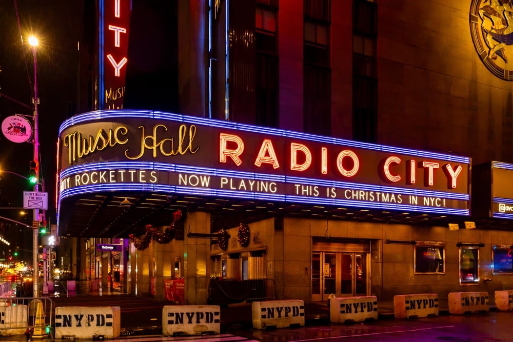 Radio City Music Hall marquee lit at night, advertising "This is Christmas in NYC" with barricades below.