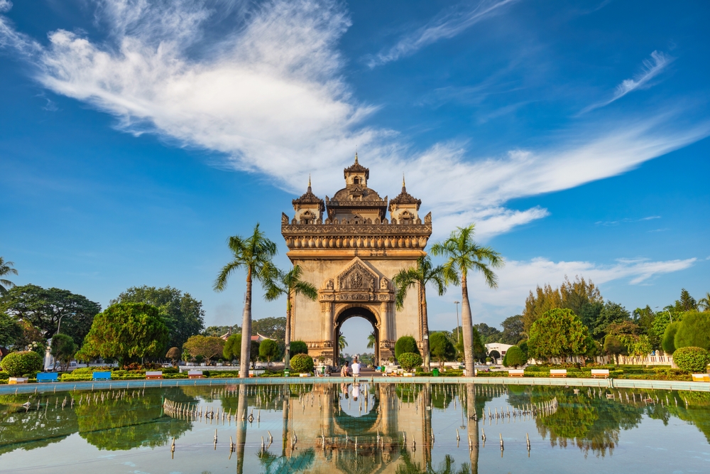Ornate arch with palm trees reflected in a pond under a blue sky with scattered clouds.