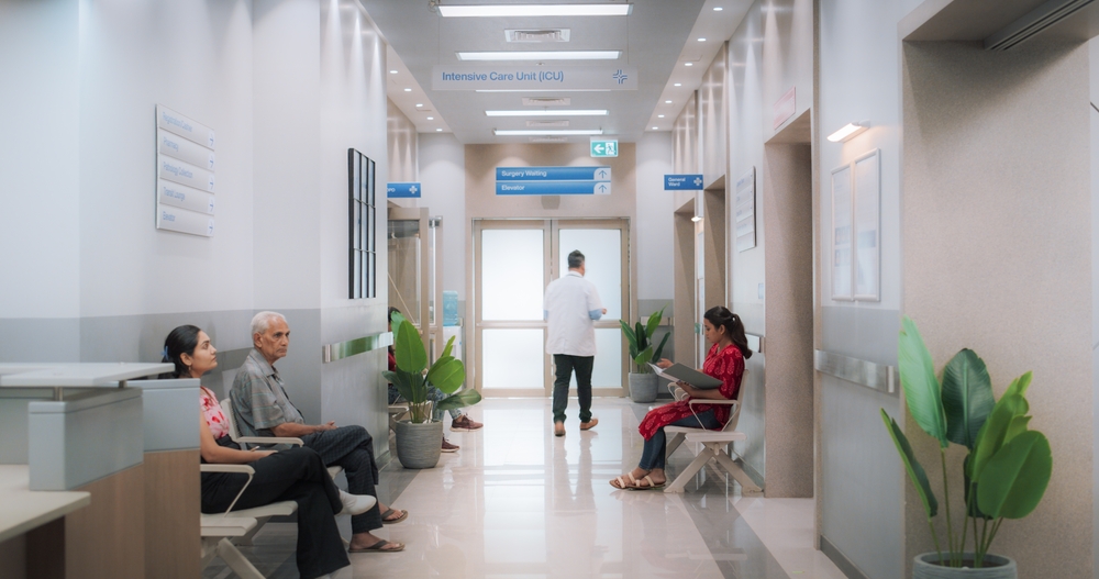 Hospital corridor with people seated, a doctor walking, and signs indicating Intensive Care Unit (ICU) direction.