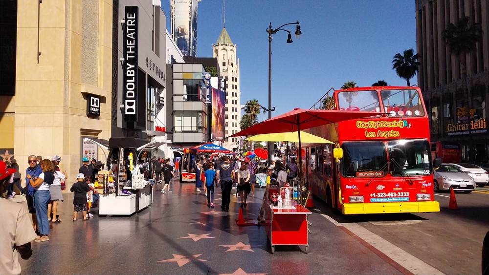 Busy street scene with red sightseeing bus, street vendors, pedestrians, and stars on the sidewalk.
