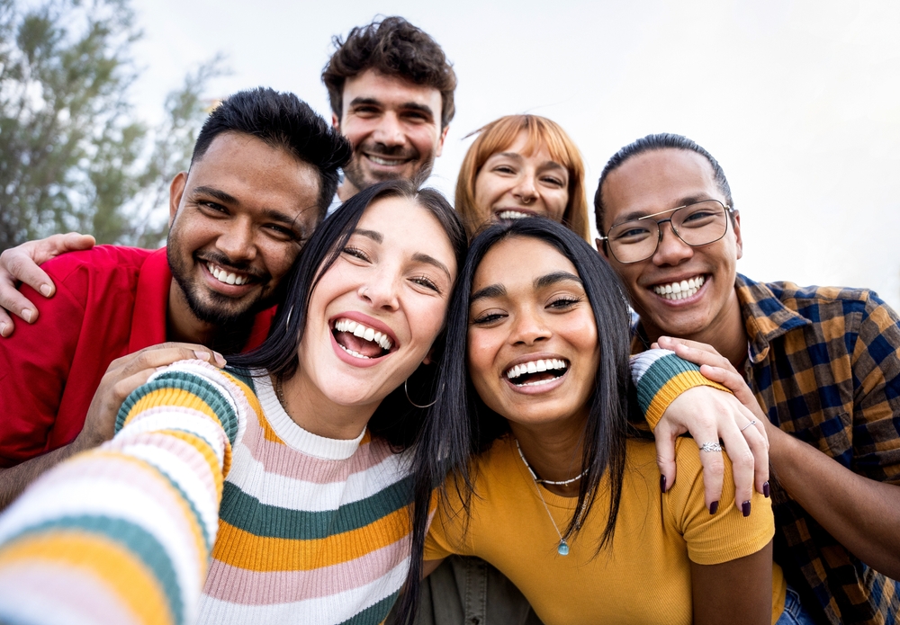 Six people smiling and taking a selfie outdoors, with trees in the background.