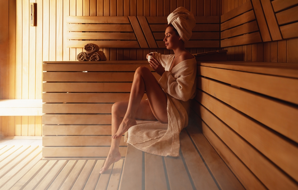 Person in towel and robe sits in wooden sauna, holding a cup and relaxing.