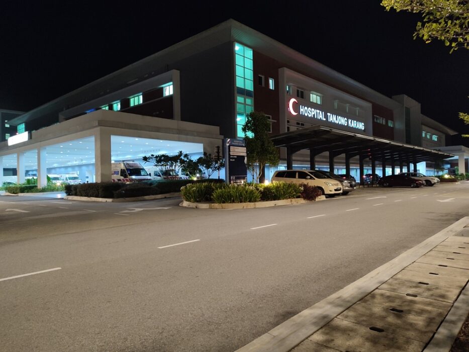 Night view of Hospital Tanjong Karang entrance with parked vehicles and ambulances under bright lights.