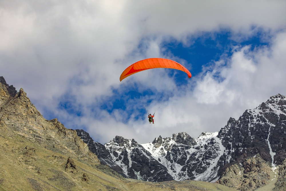 Person paragliding with red canopy over snow-capped mountains under cloudy sky.