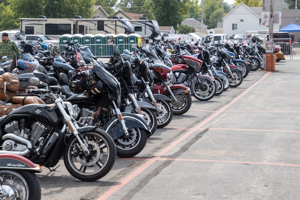 Several parked motorcycles lined up in rows on a paved lot with porta-potties in the background.