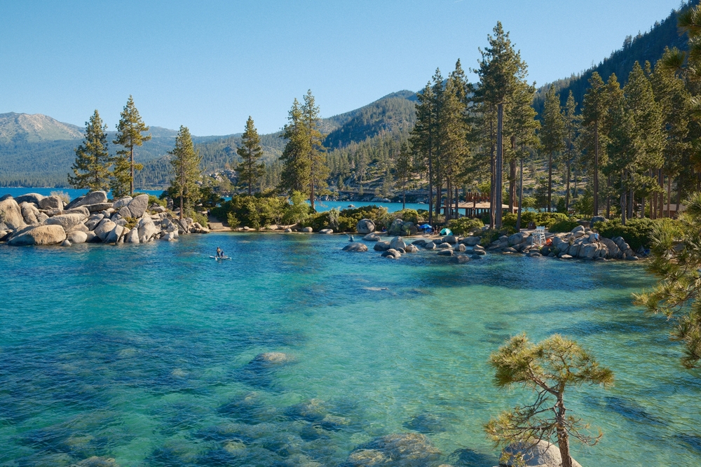 Clear blue lake with rocky shoreline, pine trees, and a person paddleboarding under a clear blue sky.