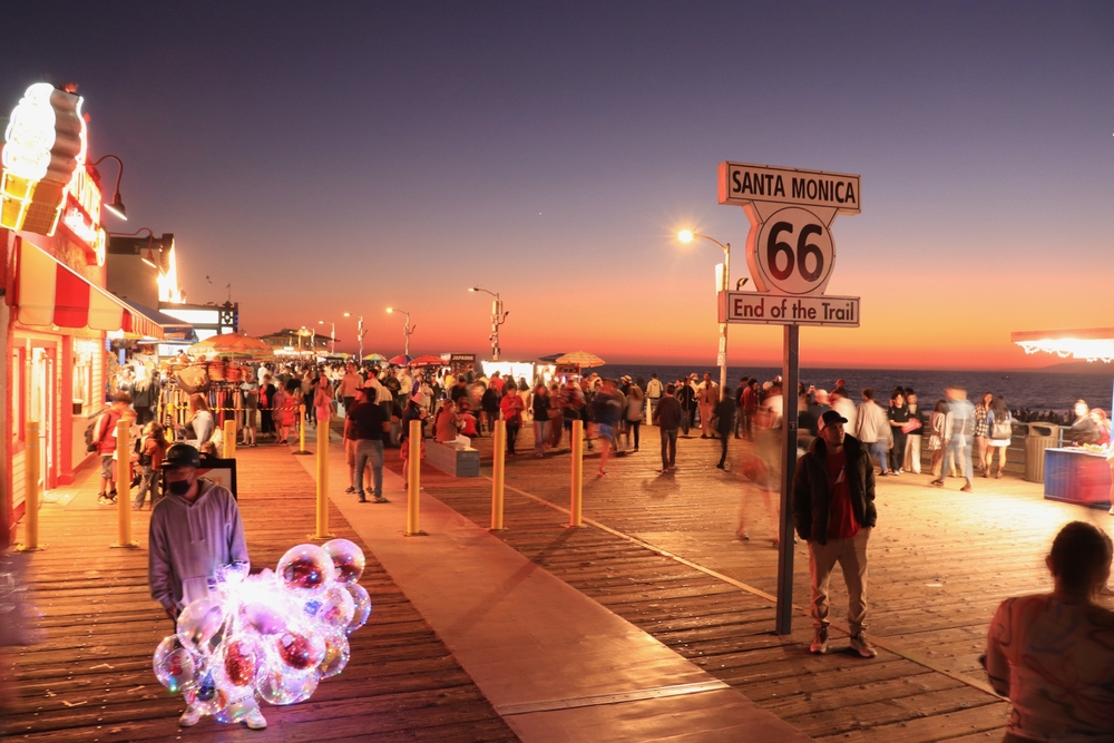 Crowded Santa Monica Pier at sunset with Route 66 sign and balloon vendor in foreground.