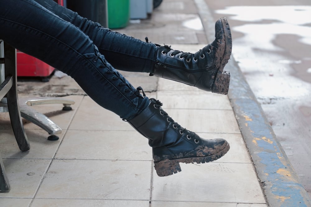Two legs wearing muddy black lace-up boots, jeans, seated on a tiled surface.