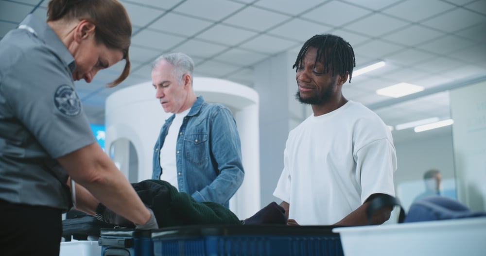 Security officer inspects luggage at airport checkpoint, two men stand by, one smiling.