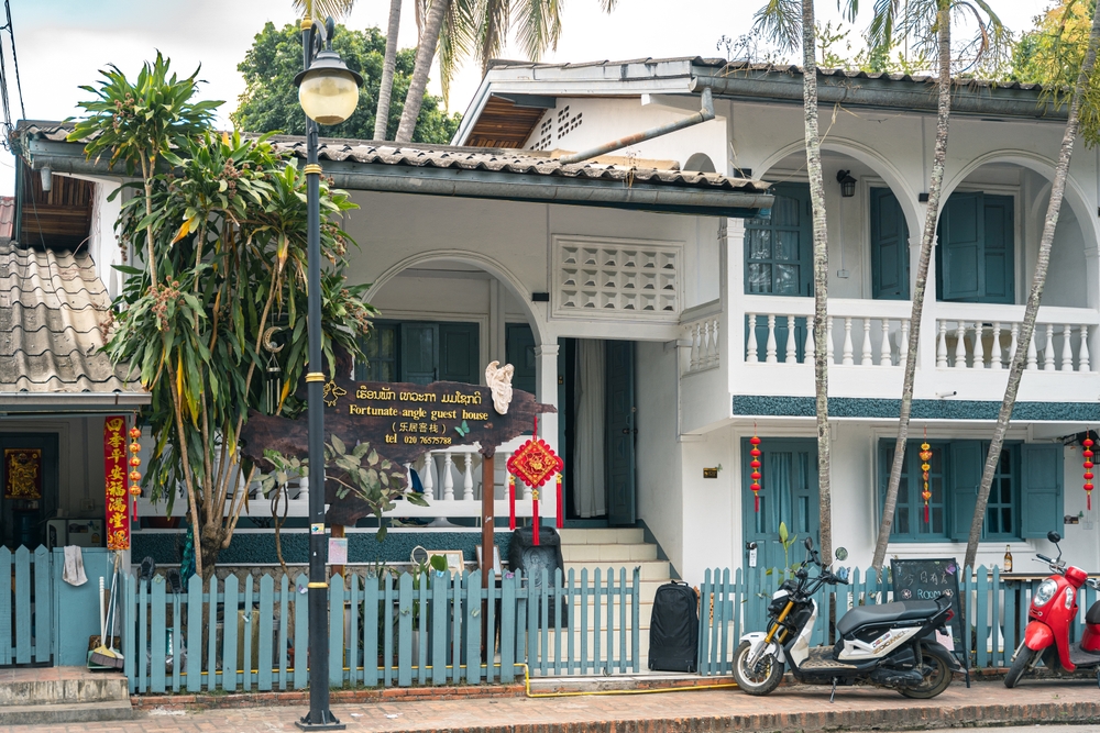 Two-story guesthouse with arches, balcony, blue shutters, sign, plants, parked motorbike, and red lantern decorations.