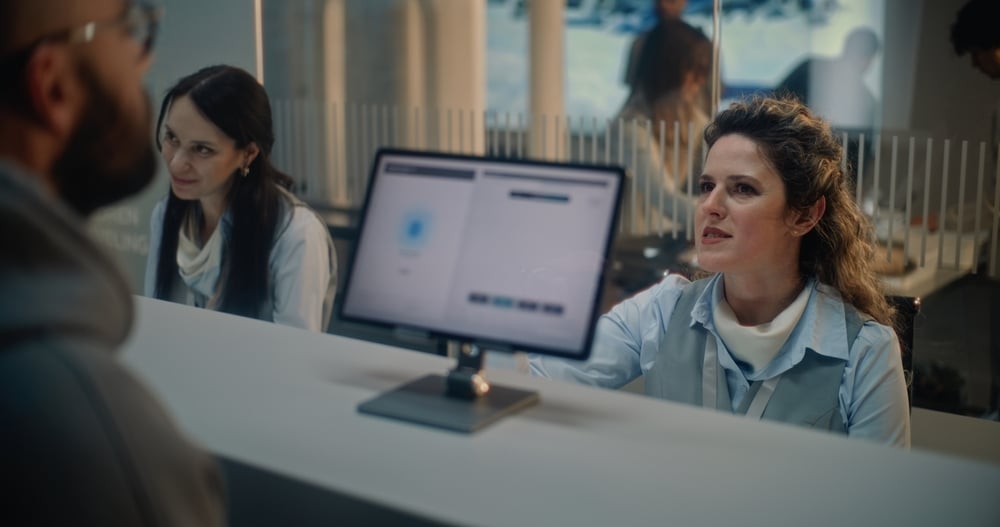 A receptionist talks to a person at a desk with a computer screen facing her.