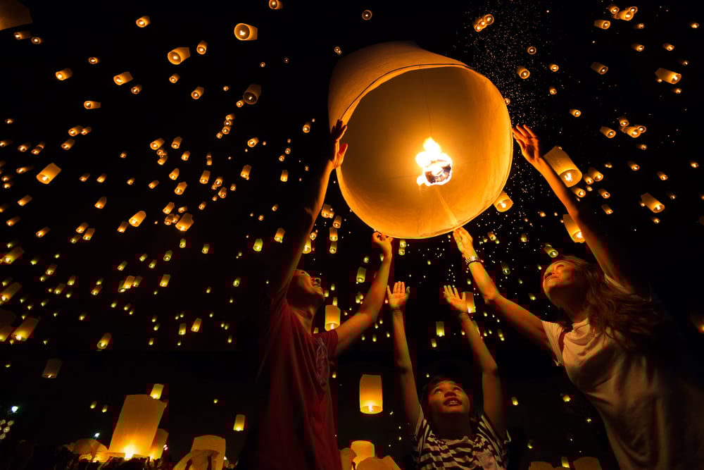 Three people releasing a lit paper lantern into the sky among many floating lanterns at night.