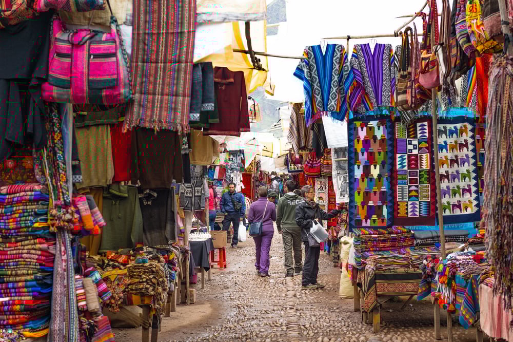 Outdoor market with colorful woven textiles displayed on both sides of a cobblestone street, people walking and browsing.