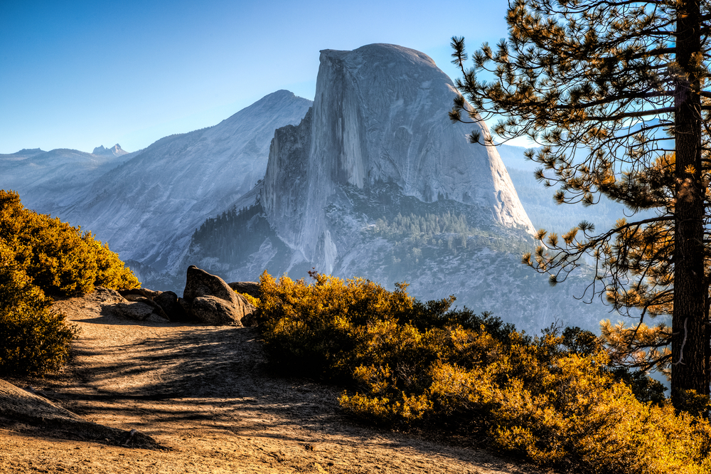A rocky path with shrubs and trees overlooks a large granite mountain under a clear blue sky.