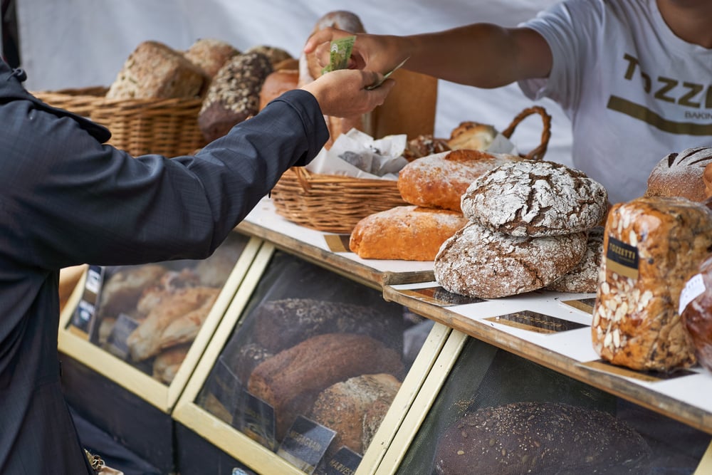 Customer purchasing bread at a market stall with variety of loaves displayed, including multigrain and rustic.