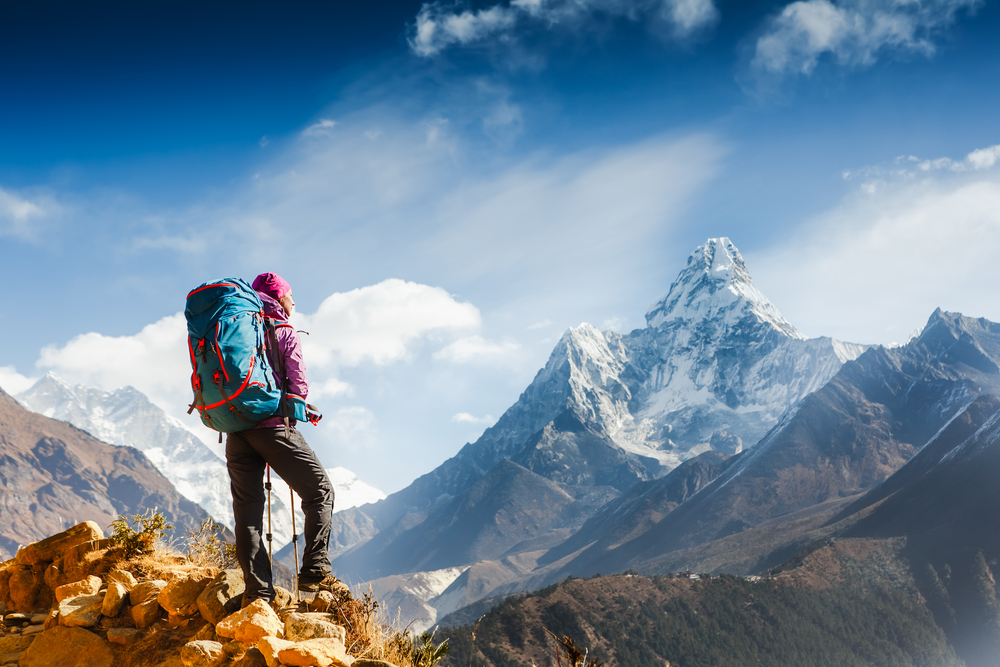 Hiker with backpack standing on rocky terrain, overlooking snow-capped mountains under a clear blue sky.