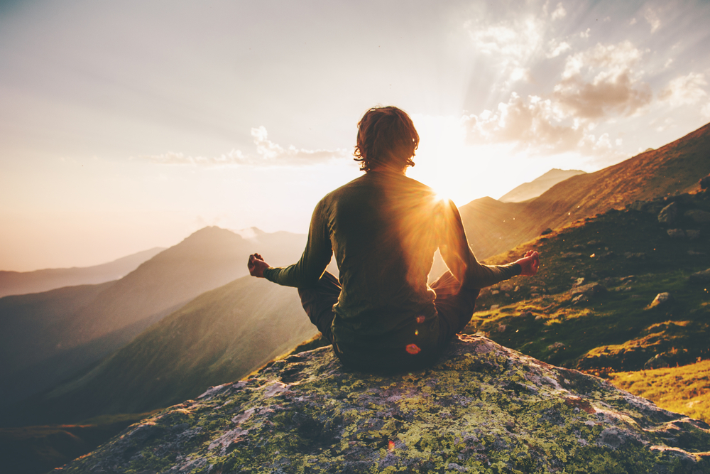 Person meditating on a rock, facing sunset over mountains with sunlight creating a silhouette effect.