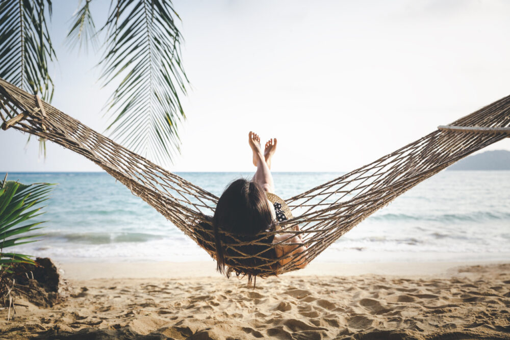 Person relaxing on hammock at beach, ocean and palm leaves in background.