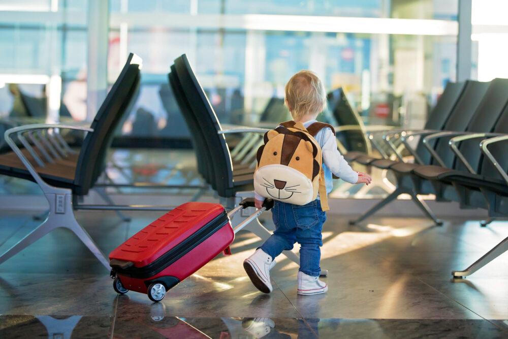 Toddler with lion backpack pulling red suitcase in airport terminal, near empty seating area.