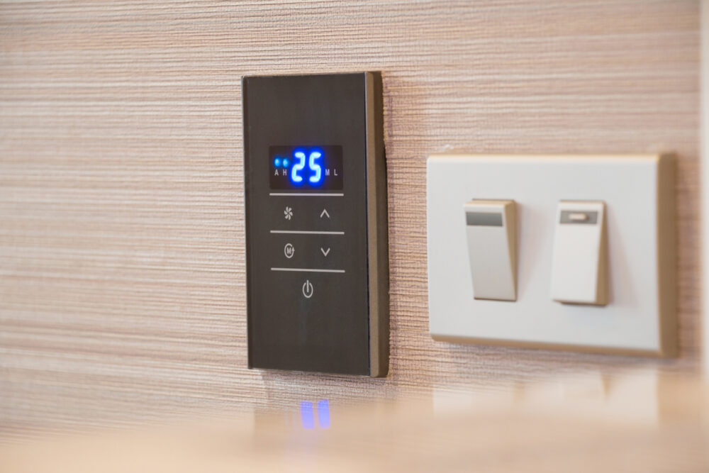 Black digital thermostat displaying 25 degrees next to two white light switches on a textured wall.