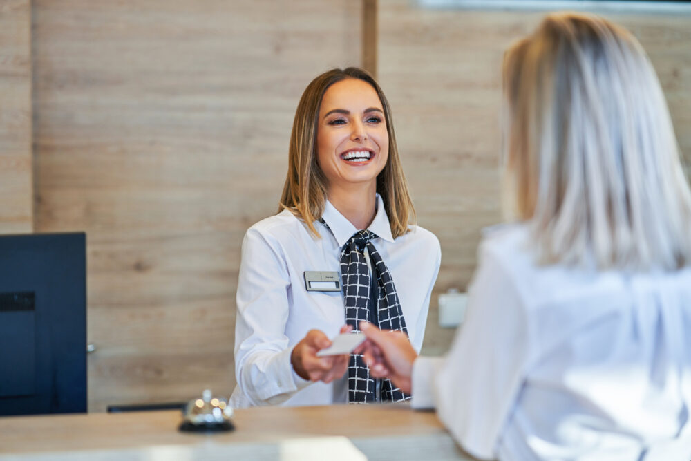Smiling receptionist in uniform hands card to customer at a hotel front desk.
