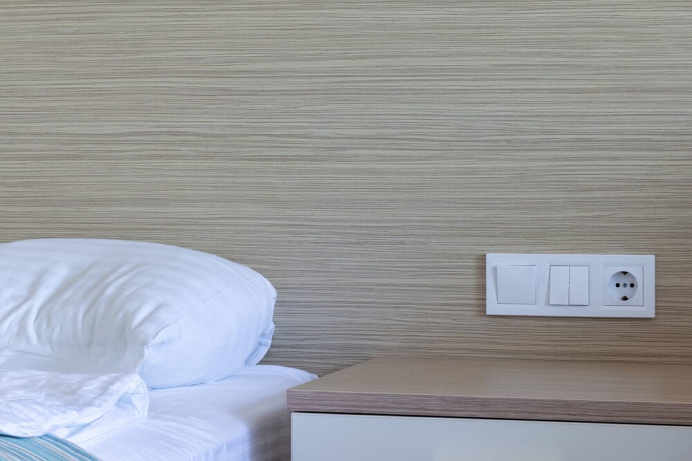 Bedroom interior with striped pillow, wooden nightstand, and wall outlets on beige wall background.
