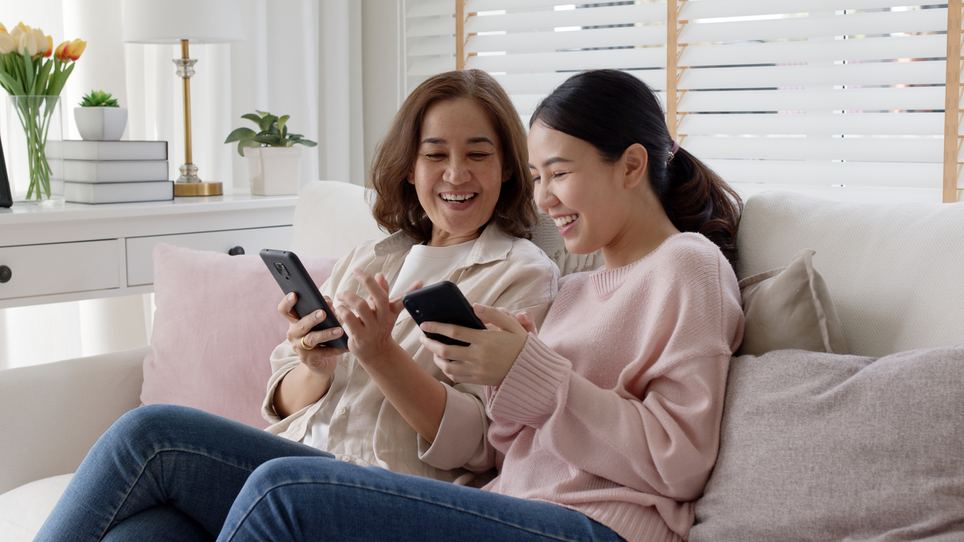Two women sitting on a couch, smiling and looking at smartphones, with a lamp and books in background.