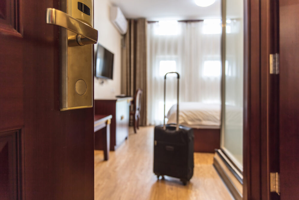 Hotel room entrance with open door, visible bed, suitcase, television, and wooden floor.