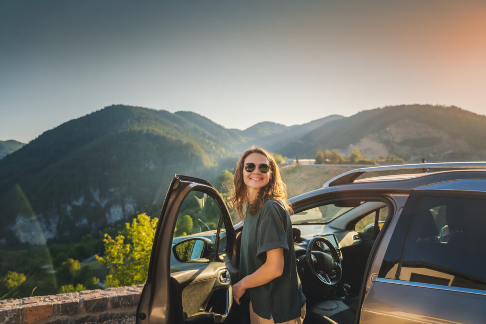 A woman with sunglasses smiling near an open car door, with mountains in the background.