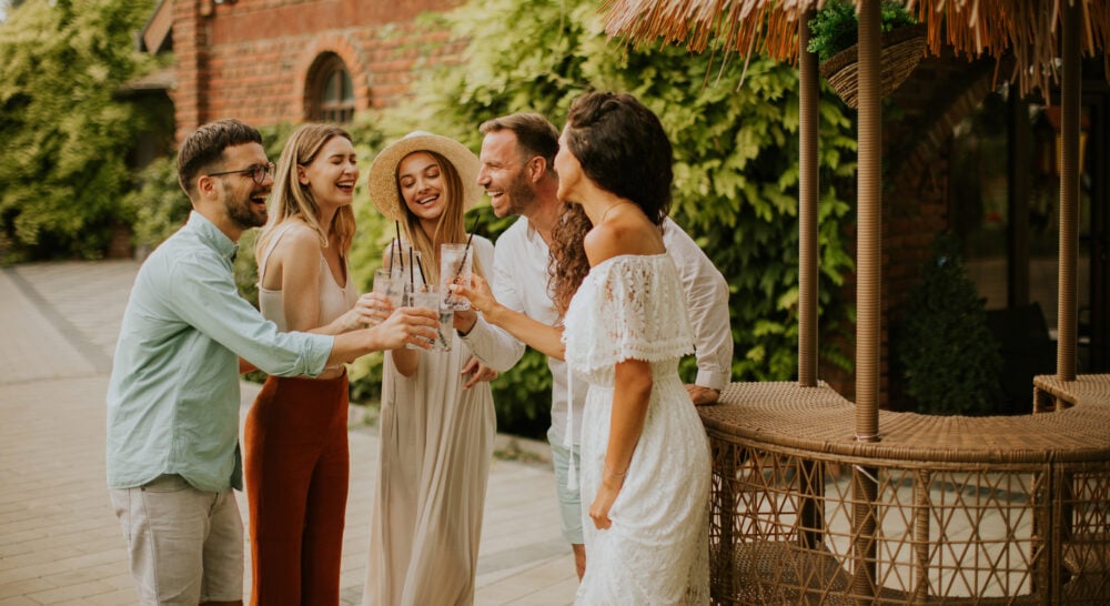 Five people smiling and toasting with drinks outside near wicker furniture and greenery.