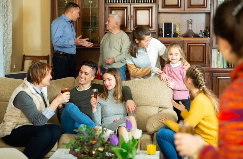 People gathered in a living room, smiling, holding drinks, and engaging in conversation.