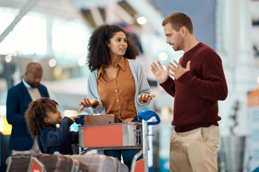 A woman and man talking at an airport, with a child and luggage cart nearby.