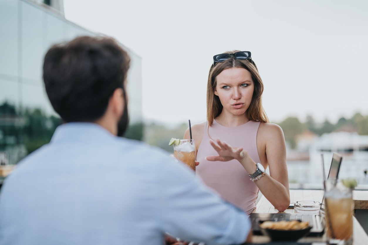 Woman gesturing while talking to a seated man at an outdoor café table.