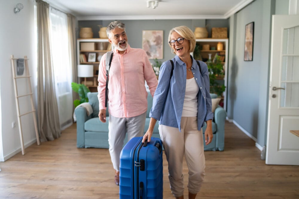 Smiling couple with backpacks entering a living room, one carrying a large blue suitcase.