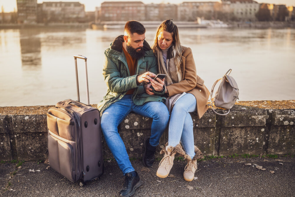 Couple sitting on a riverside ledge sharing a smartphone, with luggage and backpacks beside them.