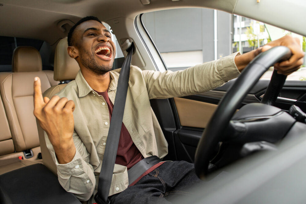 Man driving car, singing with enthusiasm, left hand raised, wearing seatbelt and casual clothes.