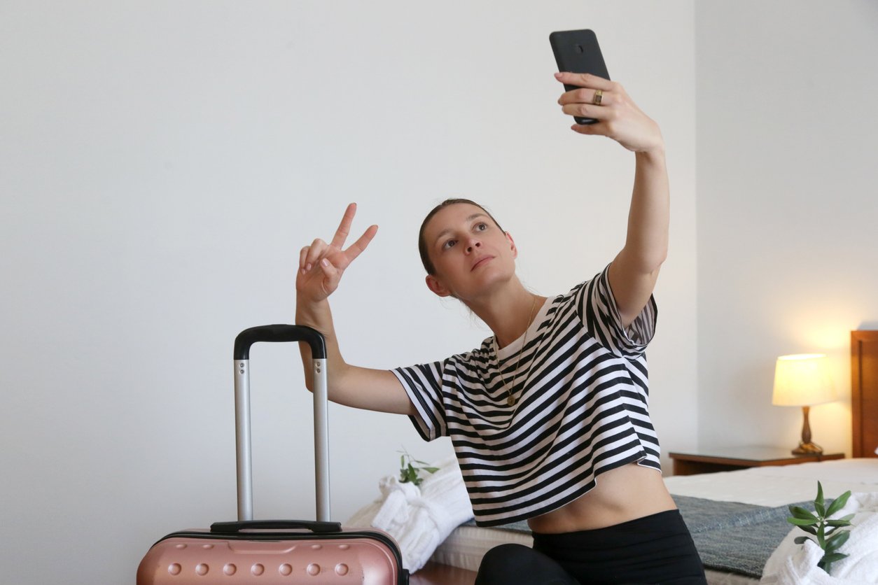 Person taking a selfie with smartphone, making peace sign, beside a pink suitcase in a bedroom.