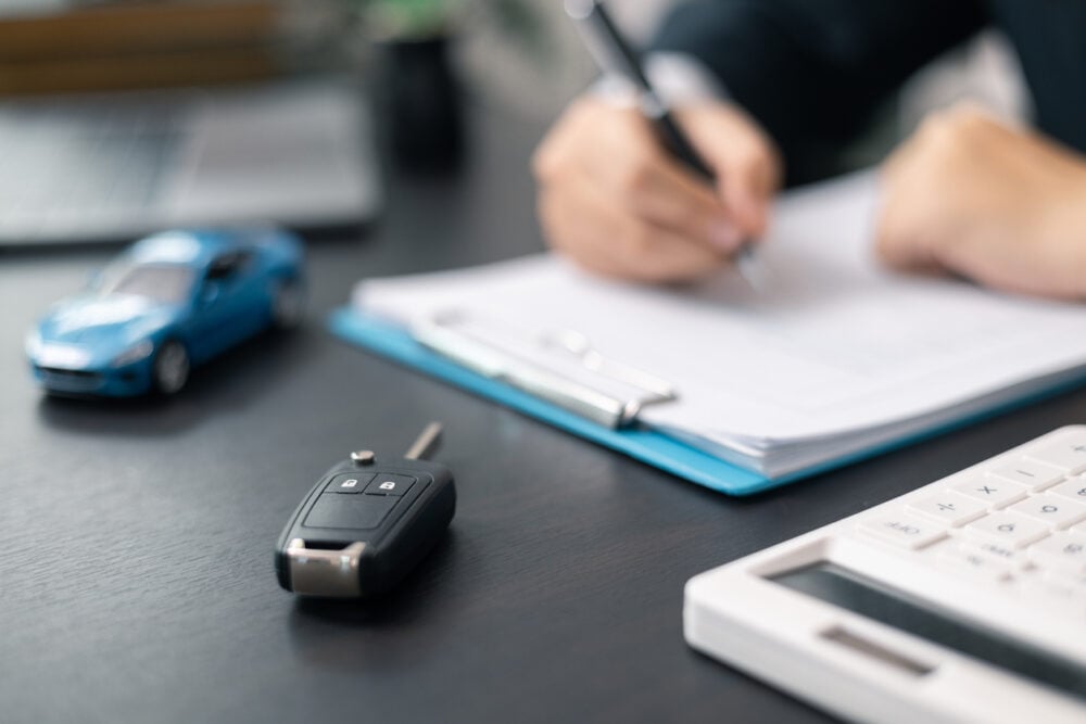 Car key, toy car, and calculator on a table with a person writing on a clipboard.