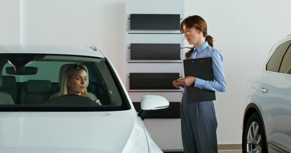 A woman in a white car talks to another woman holding a clipboard in a showroom.