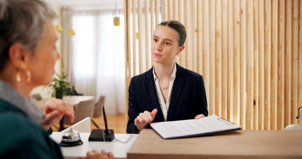 A woman in a suit holds a clipboard and talks to another woman at a reception desk.