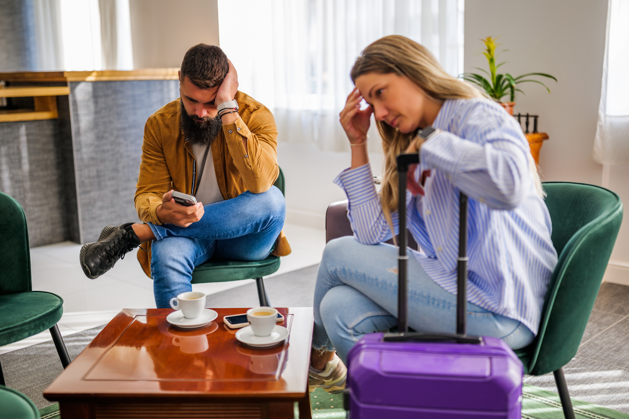 Two people seated with luggage and coffee cups, looking upset, in a bright room.