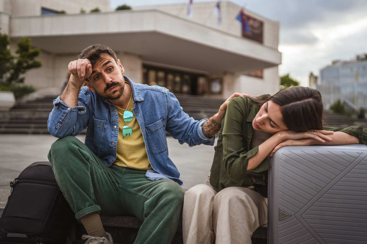 A man in a denim jacket sits next to a woman resting on luggage outside a building.