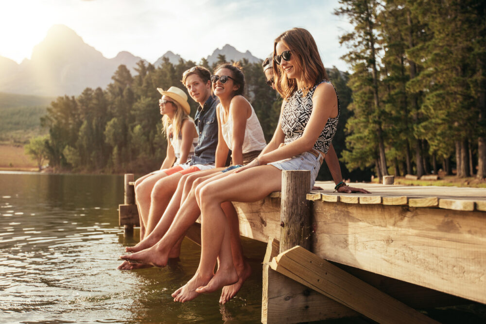 Six people sitting on a wooden dock, feet in water, with trees and mountains in the background.